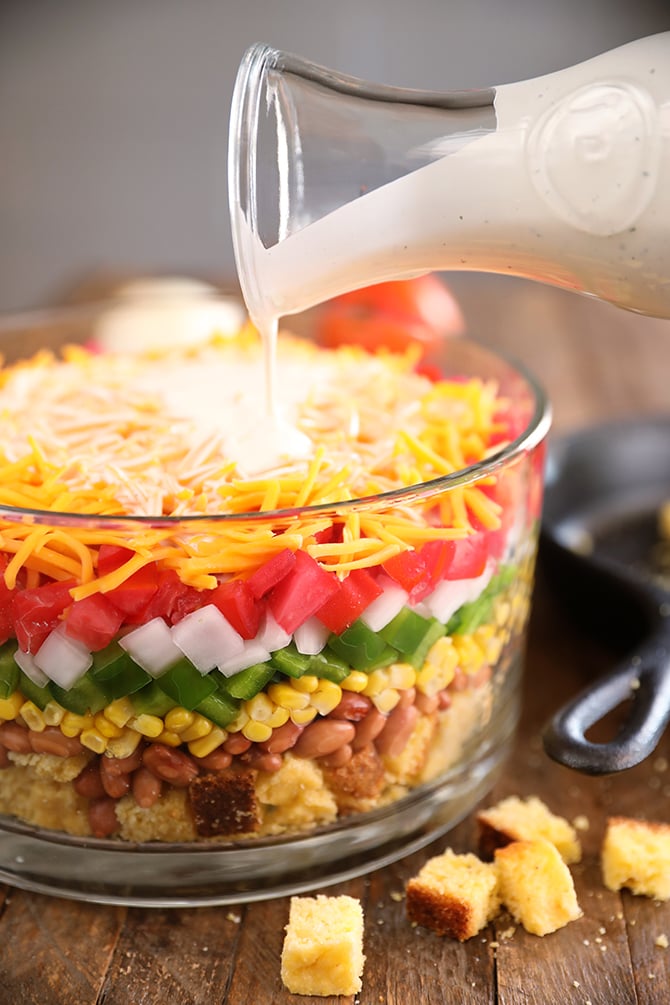 Layered Cornbread Salad in clear glass bowl - buttermilk ranch being poured over the top